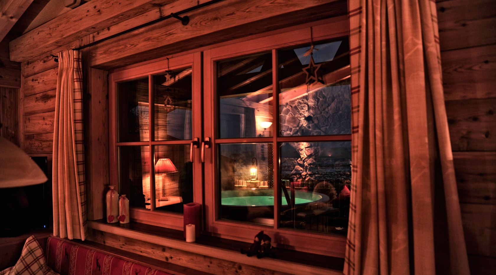 Abendlicher Blick aus dem Chaletfenster auf beleuchtete Terrasse mit Whirlpool und stimmungsvoller Lichtkulisse.
