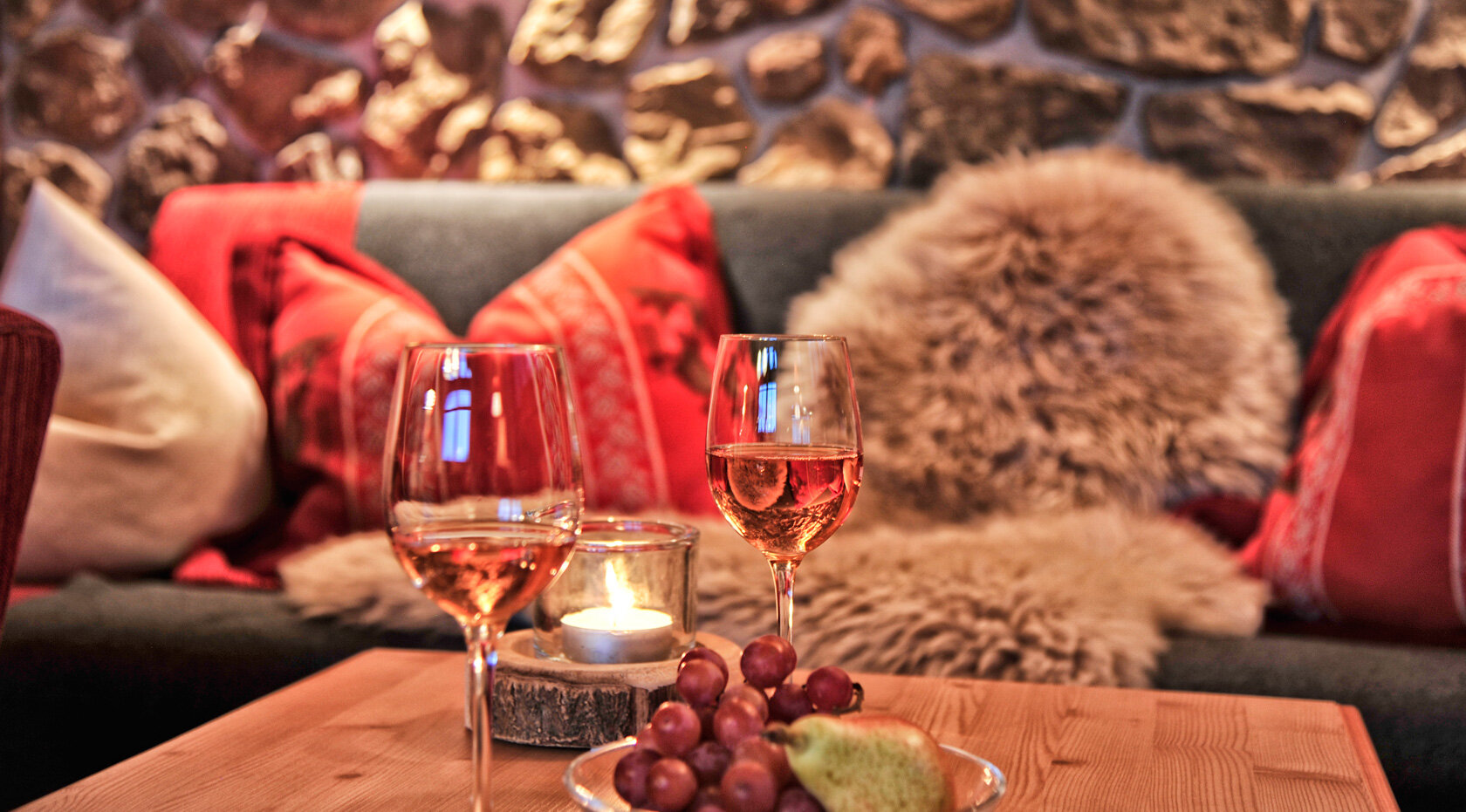 Zwei Weingläser, ein Teelicht, Trauben und eine Birne auf einem Couchtisch vor einem gemütlichen Sofa im Chalet Rustika.