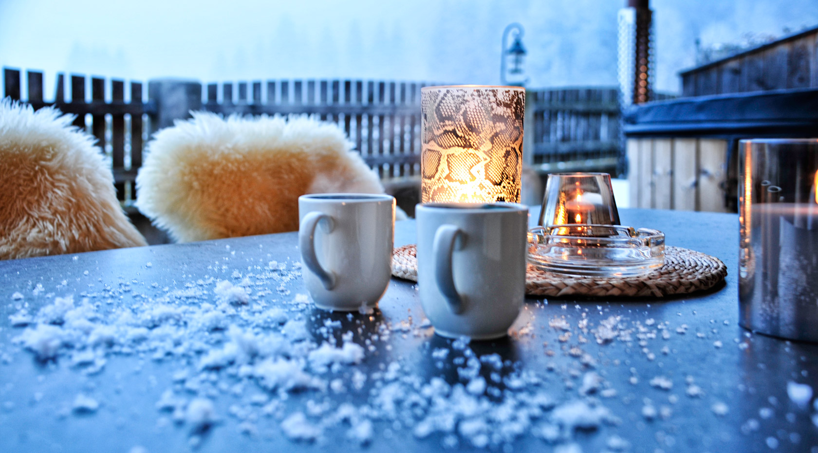 Zwei Tassen auf verschneitem Tisch mit Kerzen, Fellen und winterlichem Bergblick auf einer gemütlichen Terrasse.