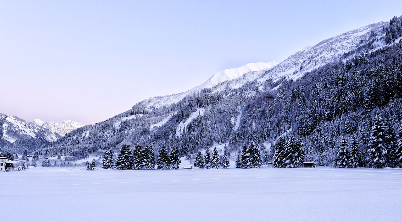 Verschneite Winterlandschaft mit Bergen, Wald und offenen Schneeflächen.