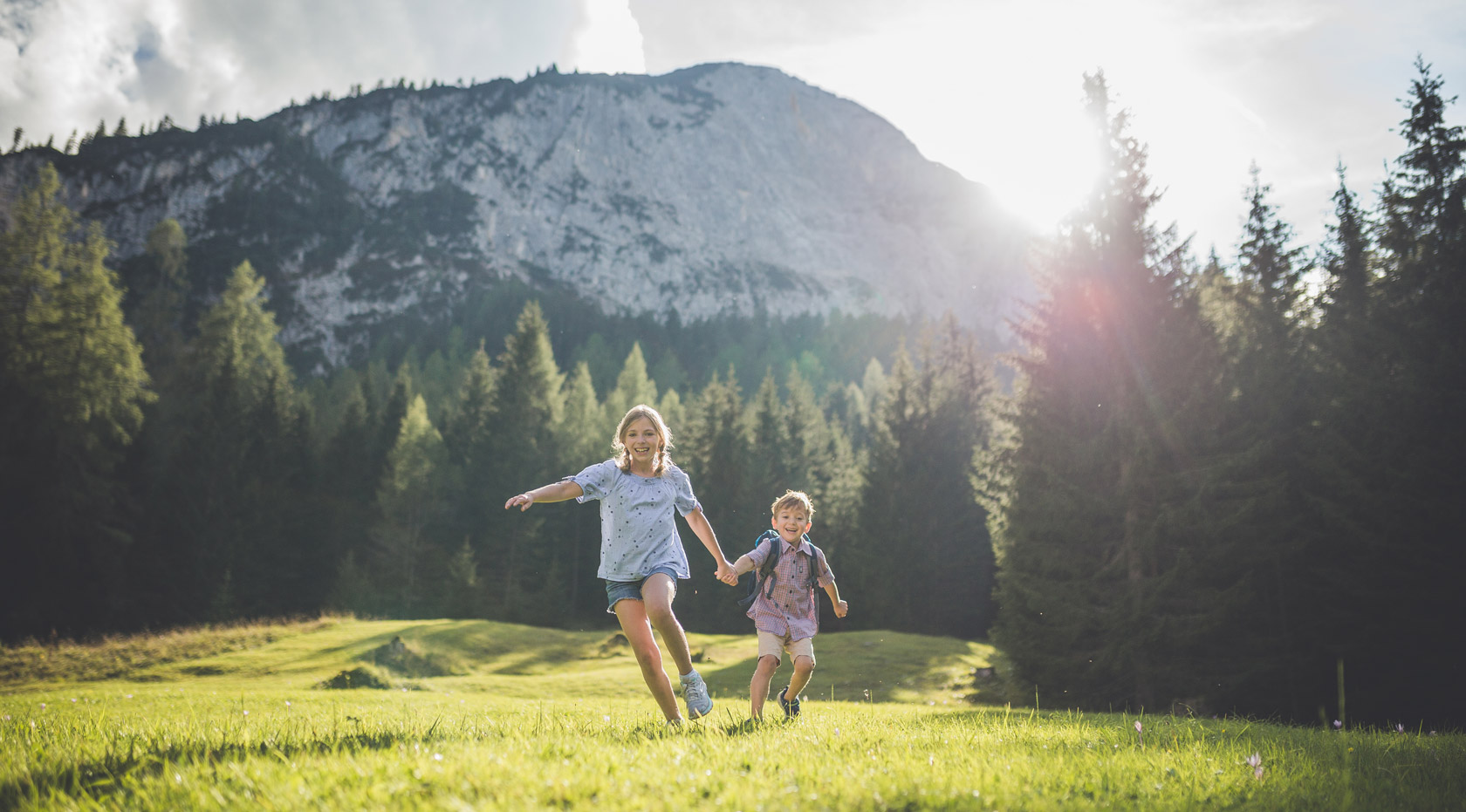 Zwei Kinder laufen lachend über eine grüne Wiese in der Morgensonne.