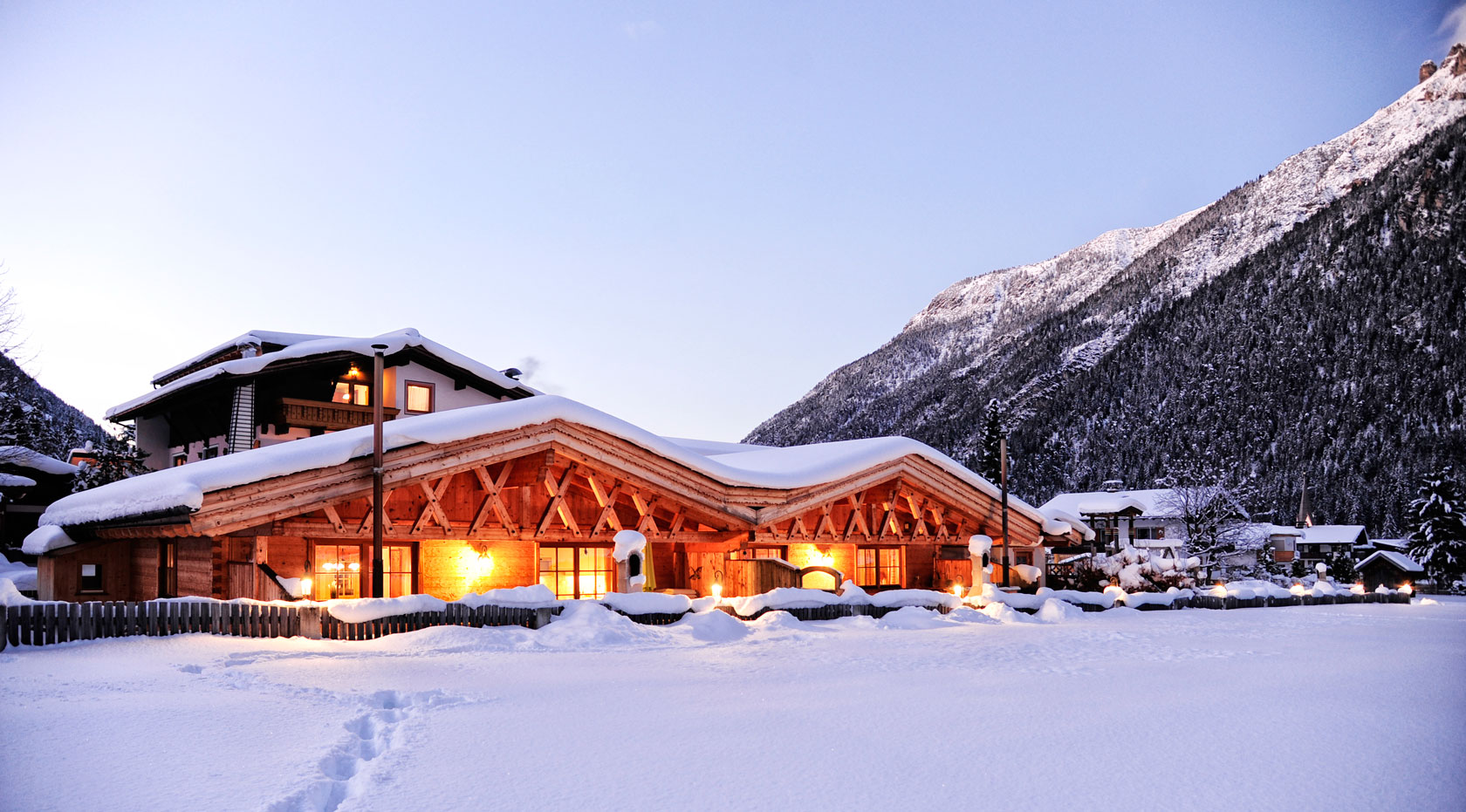 Verschneite Chalets in den Alpen bei Dämmerung, warm erleuchtet vor winterlicher Bergkulisse.